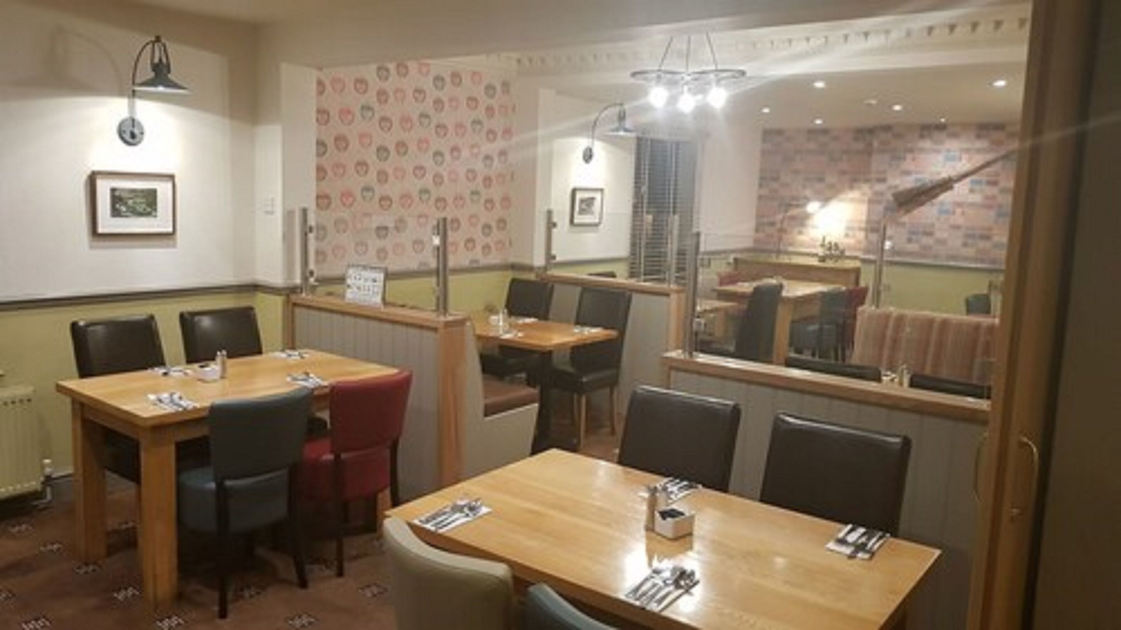 A dining area with wooden tables and chairs, a patterned wall, and a view of the kitchen through a glass partition.