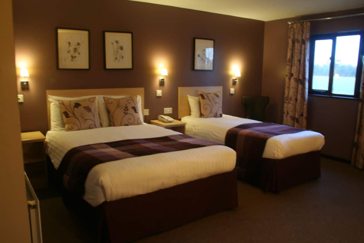 A hotel room with two beds, each with a headboard and bedside tables. There are wall-mounted lamps and framed pictures on the walls.