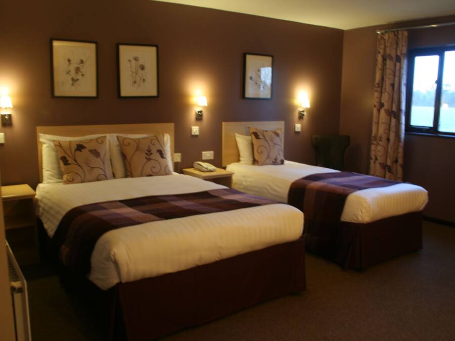A hotel room with two beds, each with a headboard and bedside tables. There are wall-mounted lamps and framed pictures on the walls.