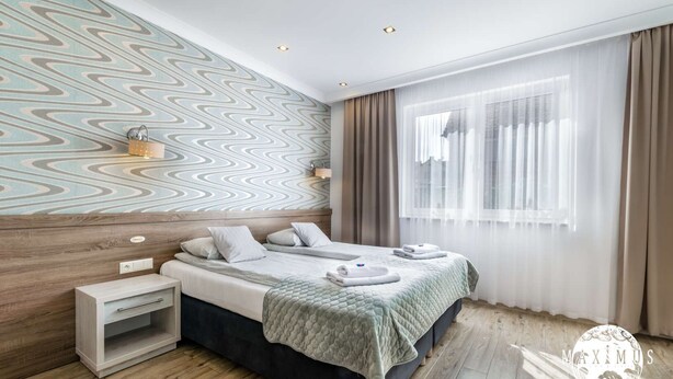 Moderni hotellihuone, jossa on suuri sänky, yöpöytä, seinään kiinnitetty valaisin ja ikkuna, jossa on ohuet verhot.