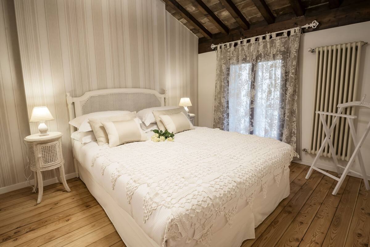 A neatly made bed with a white bedspread and pillows, a bedside table with a lamp, and a radiator on the wall.