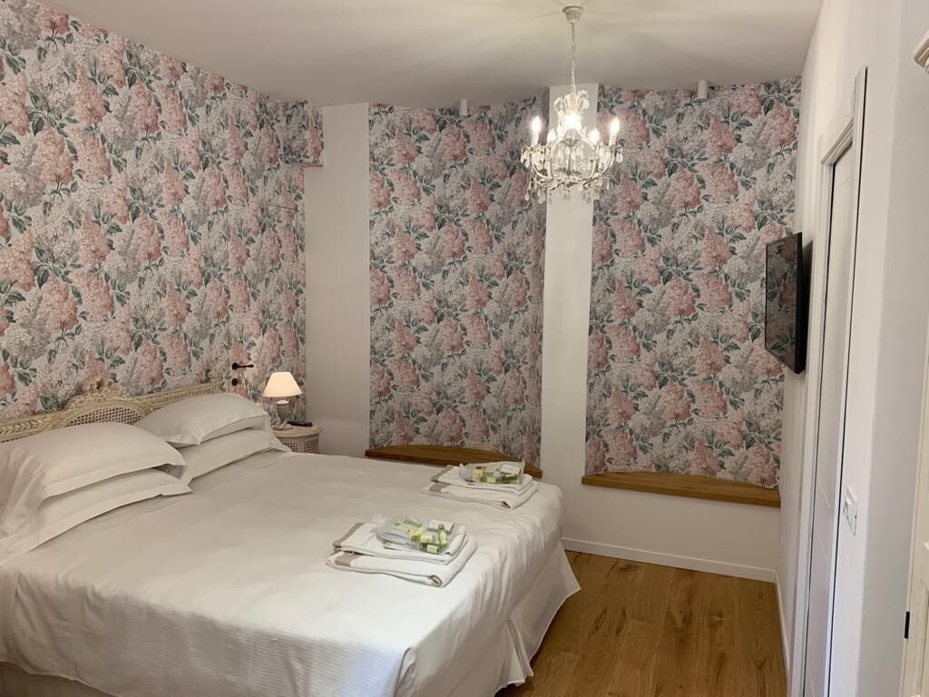 A bedroom with floral wallpaper, a chandelier, a bed with pillows and a bedside table with a lamp.
