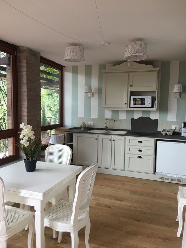 Una cocina con armarios blancos, microondas y un lavamanos. Hay una mesa de comedor con sillas y una planta en maceta.