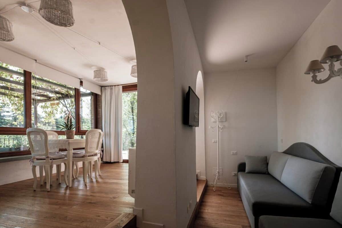 A modern living room with a wooden floor, a grey sofa, a dining table with chairs, and a TV mounted on the wall.
