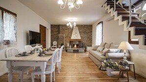 Uma sala de estar espaçosa com lareira, piso de madeira, área de jantar com cadeiras brancas e uma escada.