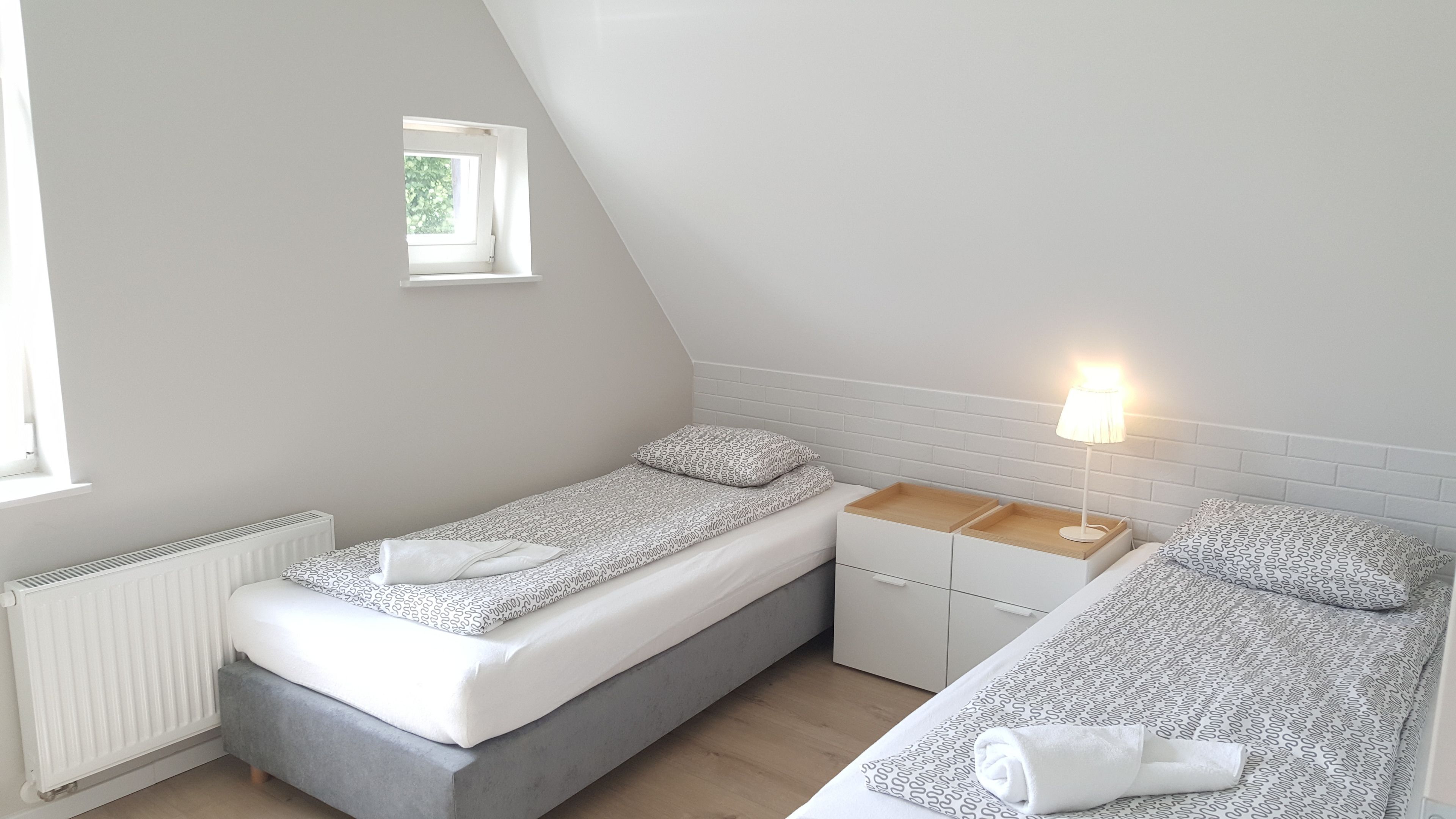 Une petite chambre à coucher moderne avec un plafond en pente, une fenêtre, un radiateur, une table de chevet avec une lampe et deux lits avec une literie à motifs.