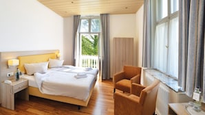 Ein Hotelzimmer mit Bett, Nachttisch, Lampe, Sessel und Blick auf Grünfläche.