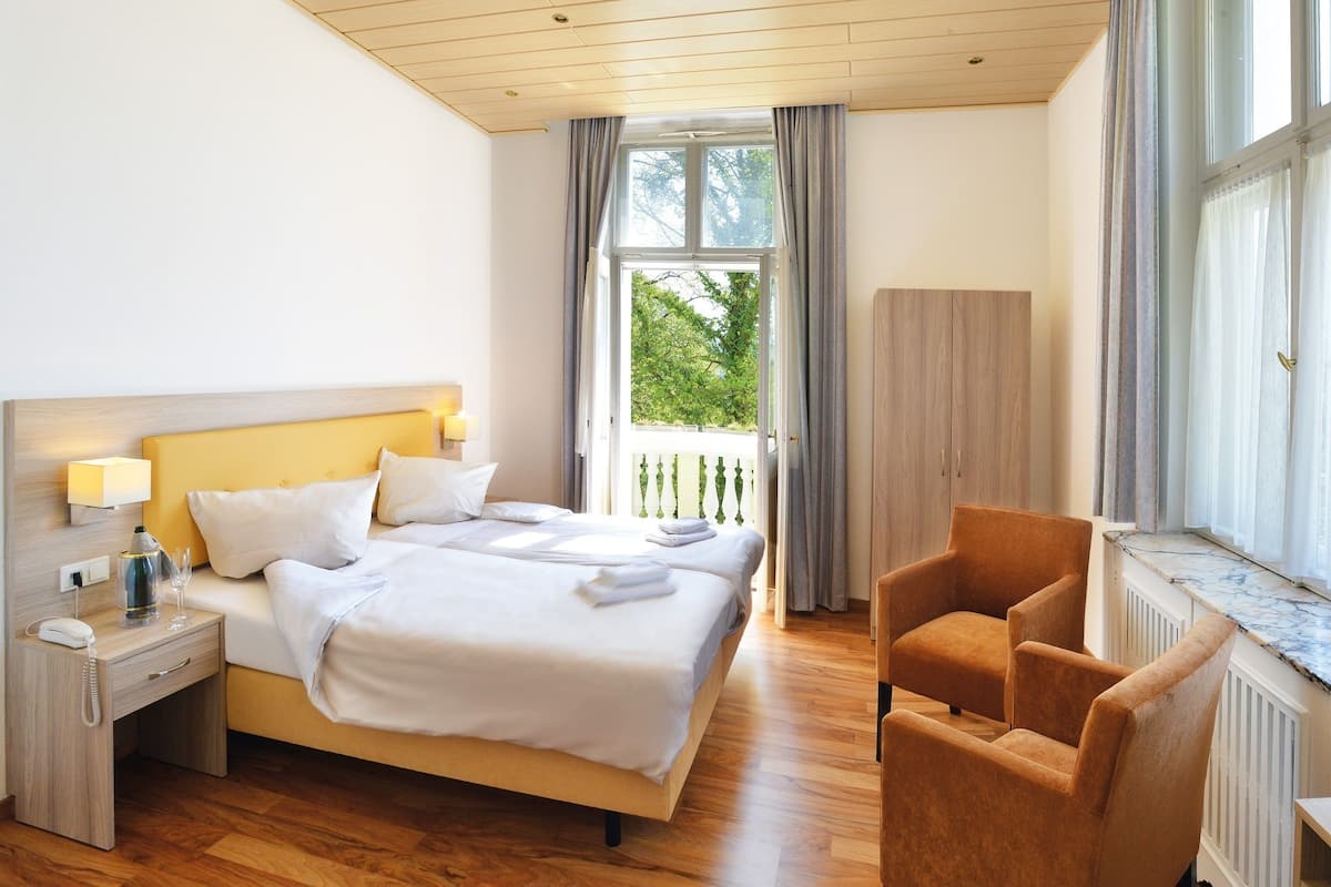 Ein Hotelzimmer mit Bett, Nachttisch, Lampe, Sessel und Blick auf Grünfläche.