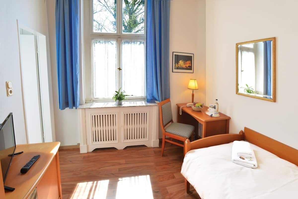 Ein Hotelzimmer mit einem Bett, einem Schreibtisch mit Lampe, einem Sessel, einem Heizkörper, einem Fenster mit Vorhängen, einem gerahmten Bild und einem Spiegel.