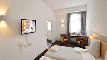 A hotel room with a bed, two armchairs, a desk with a lamp, a TV mounted on the wall, and a window with curtains.
