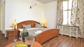 A hotel room with a wooden bed, two bedside lamps, a radiator, a window with floral curtains, and a table with fruit and a bottle.