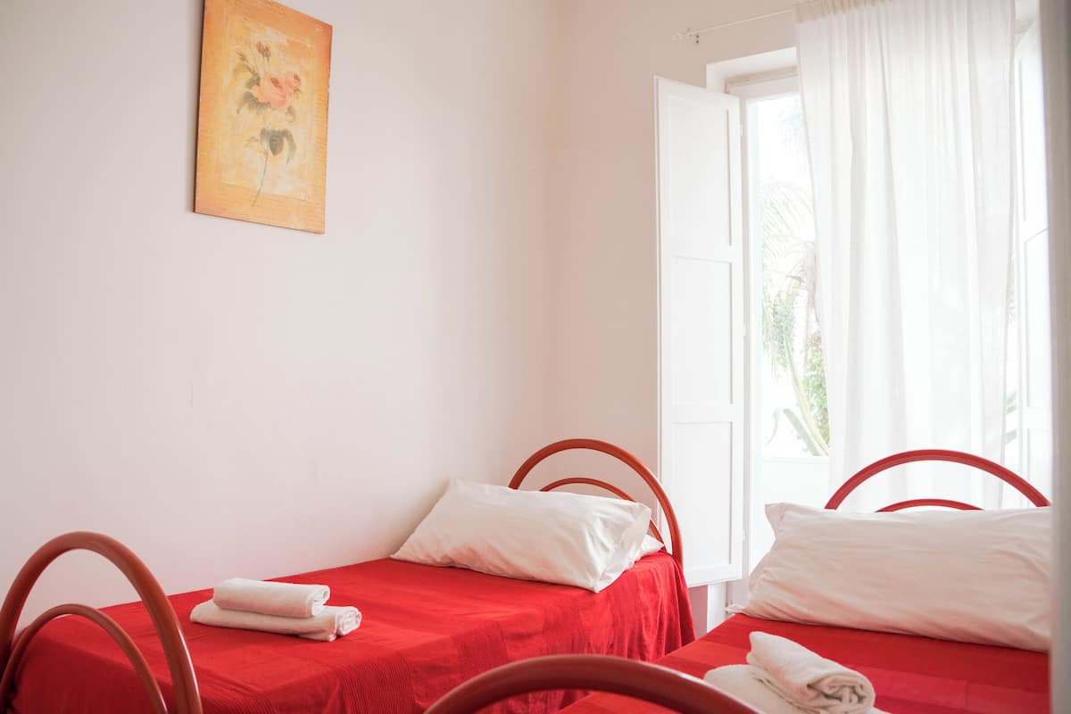Habitación con dos camas individuales, cada una con una colcha roja y almohadas blancas. En la pared hay un cuadro, una ventana con cortinas y una silla en primer plano.