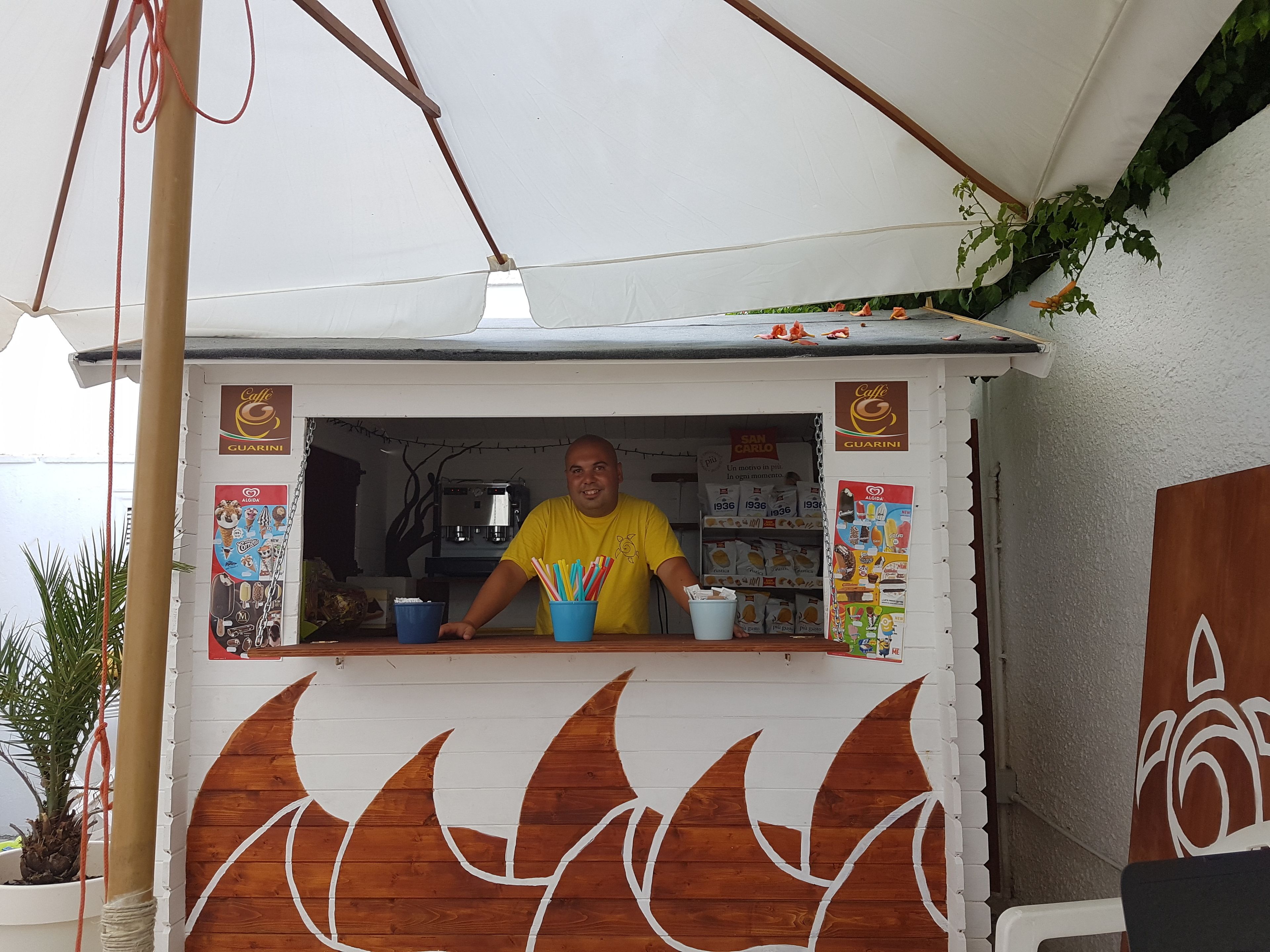 A food stall with a person behind the counter, surrounded by various food items and promotional posters.