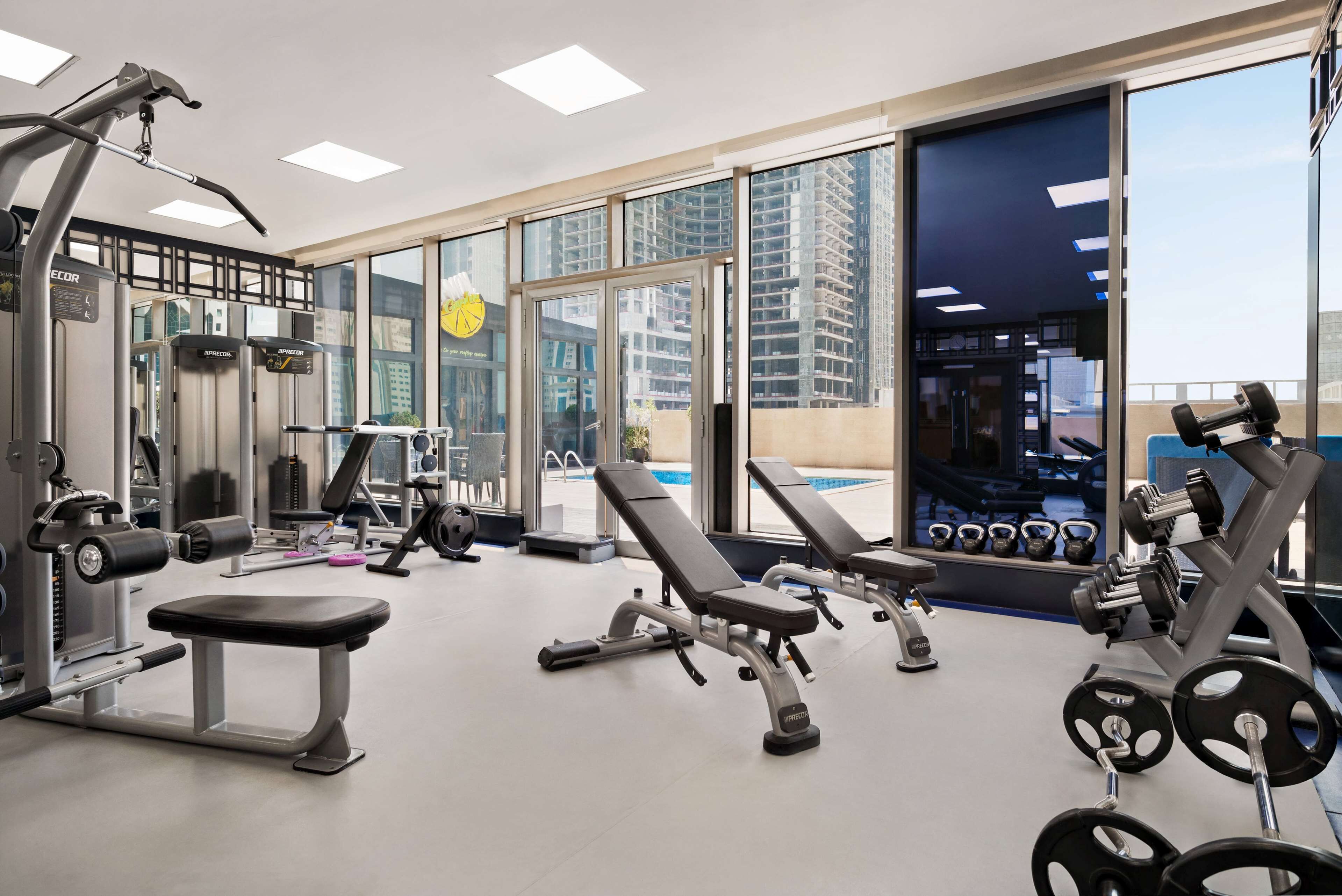 Un gimnasio moderno con diversos equipos de ejercicio, amplias ventanas y vistas a un paisaje urbano.