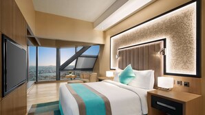 A modern hotel room with a large bed, a nightstand, a desk, a chair, and a large window offering a city view. - Wyndham Doha West Bay (Doha)