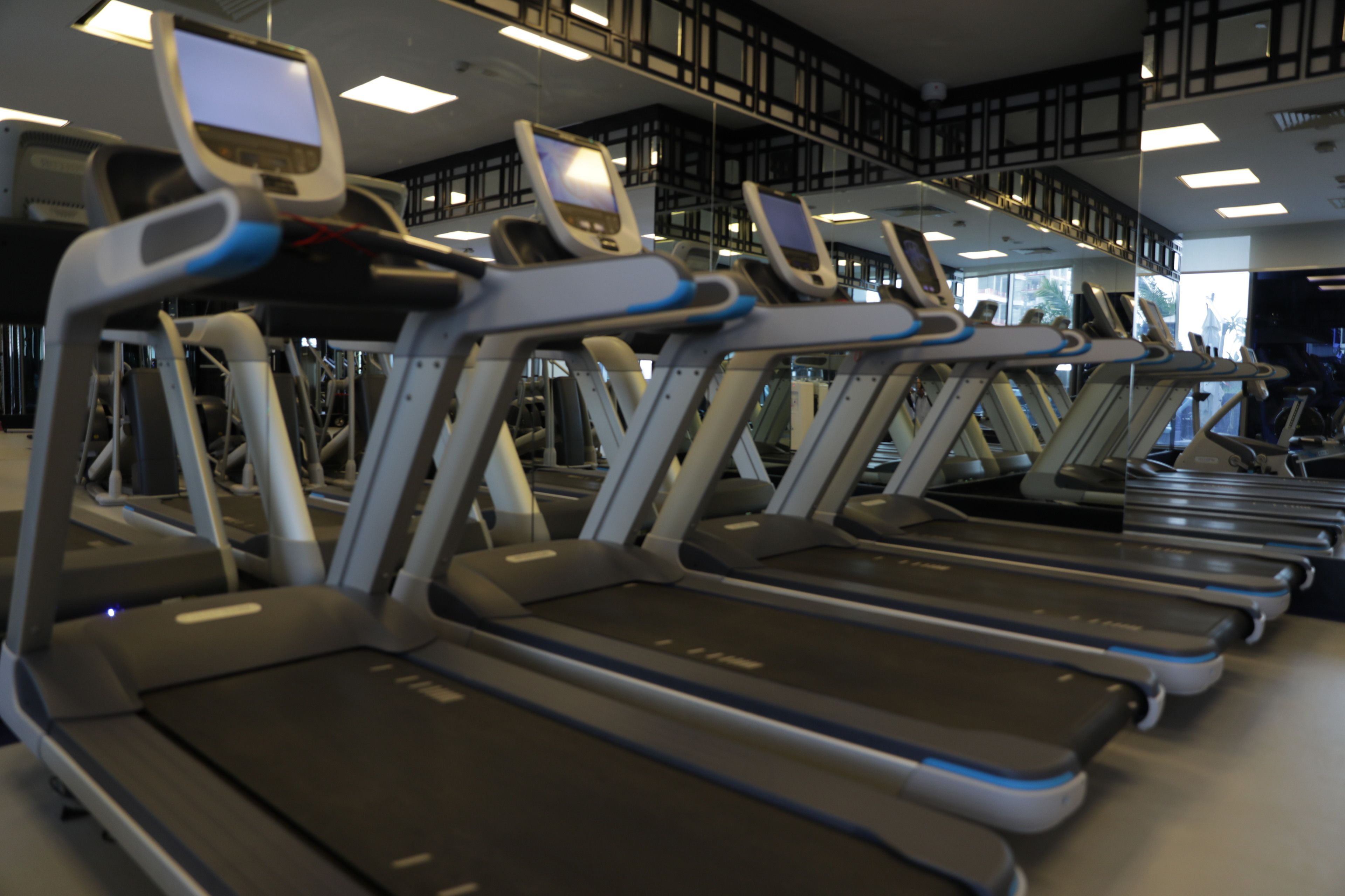 A row of treadmills in a gym with overhead lighting and a high ceiling.