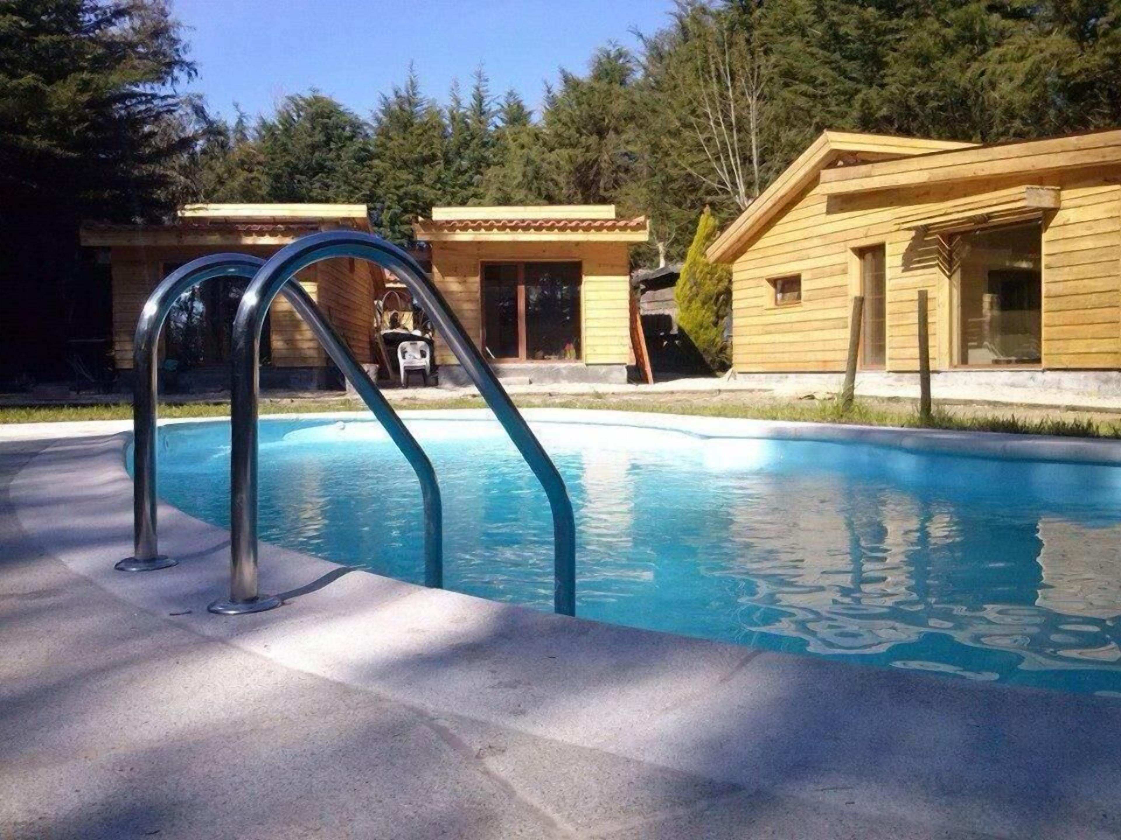 Une piscine avec des échelles en métal, entourée de bâtiments en bois et d’arbres.