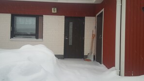 A building entrance with a black door, a window with blinds, and a number '6' above the door. Snow is piled in front of the door.
