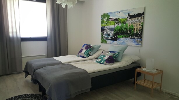 Un dormitorio con cama, mesita de noche, un cuadro y ventana con cortinas.