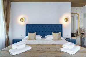 A neatly made bed with white linens and two rolled towels, a blue tufted headboard, and two wall sconces with round, gold-colored lamps. - Luxury Rooms Floramye - Adults Only (Split)