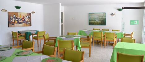 A dining area with multiple tables and chairs, green tablecloths, and a painting on the wall.