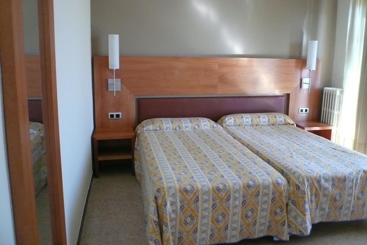 A hotel room with two beds, wooden headboard, nightstands, wall-mounted lamps, and a radiator.
