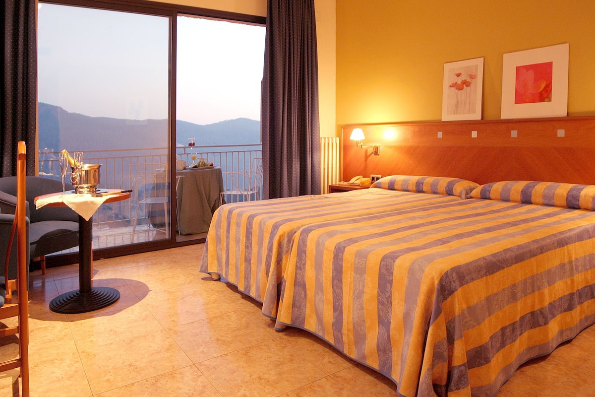 A hotel room with a bed, a desk with a chair, a table, and a view of the mountains.