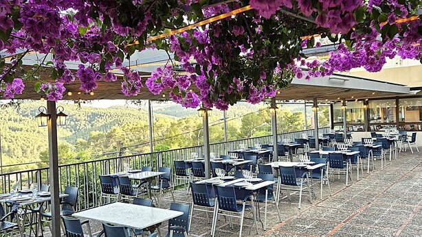 Un área de estar al aire libre con mesas y sillas, cubierta por una pérgola adornada con flores moradas, con vistas a un paisaje pintoresco.