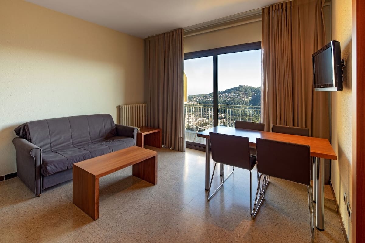 A modern hotel room with a grey sofa, wooden coffee table, and dining table with chairs. A TV is mounted on the wall, and the room has a large window with a city view.