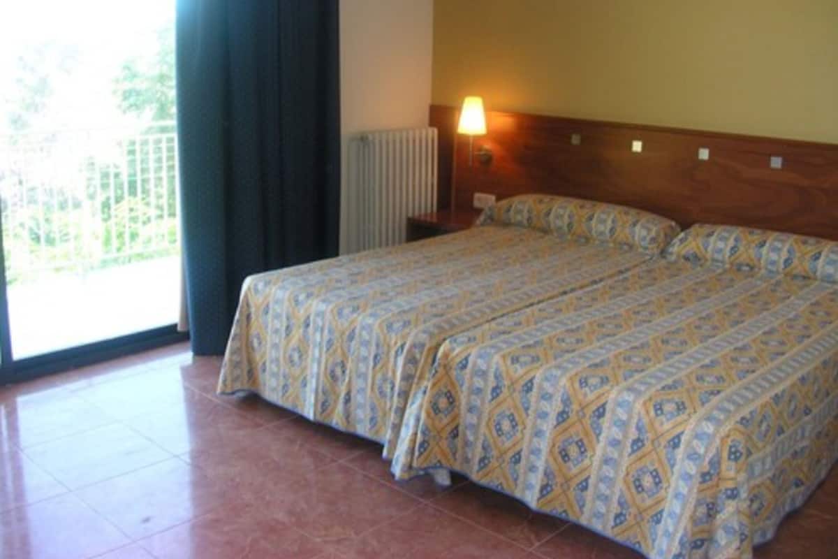 A double bed with a patterned bedspread, a nightstand with a lamp, and a view of greenery through a window.