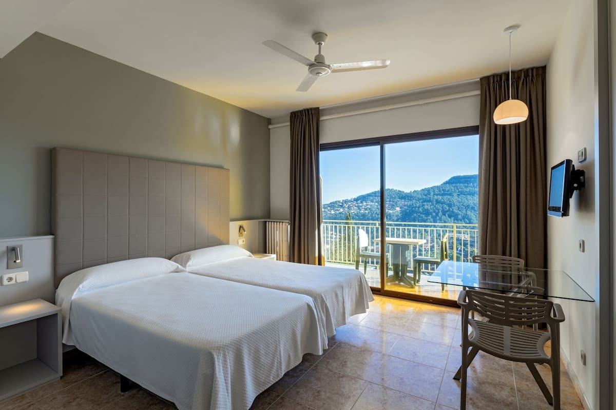 A hotel room with two beds, a ceiling fan, a desk with a chair, a TV, and a balcony with a view of the hills.