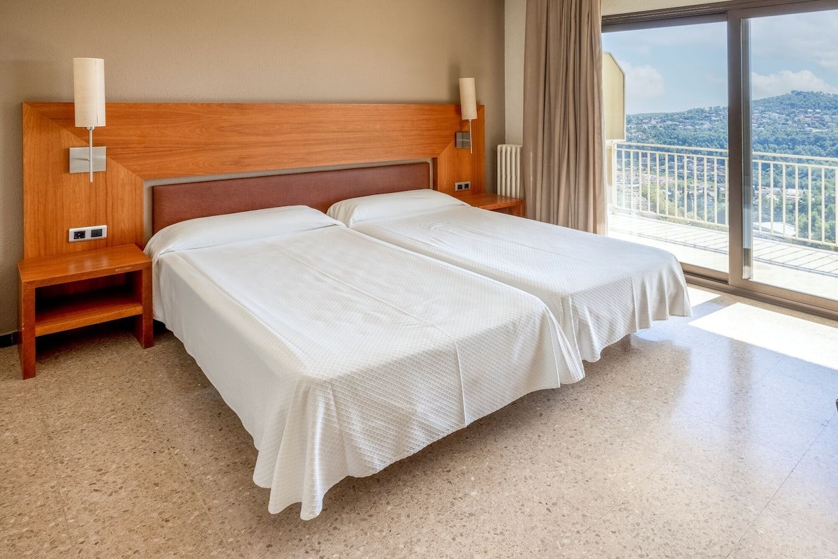 A double bed with white linens in a hotel room with a wooden headboard, bedside lamps, and a view of a balcony.