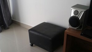 A small black ottoman with wheels, a wooden side table with a speaker, and a black chair in a room with tiled flooring. - Apartamento 3 quartos Bueno (Goiania)