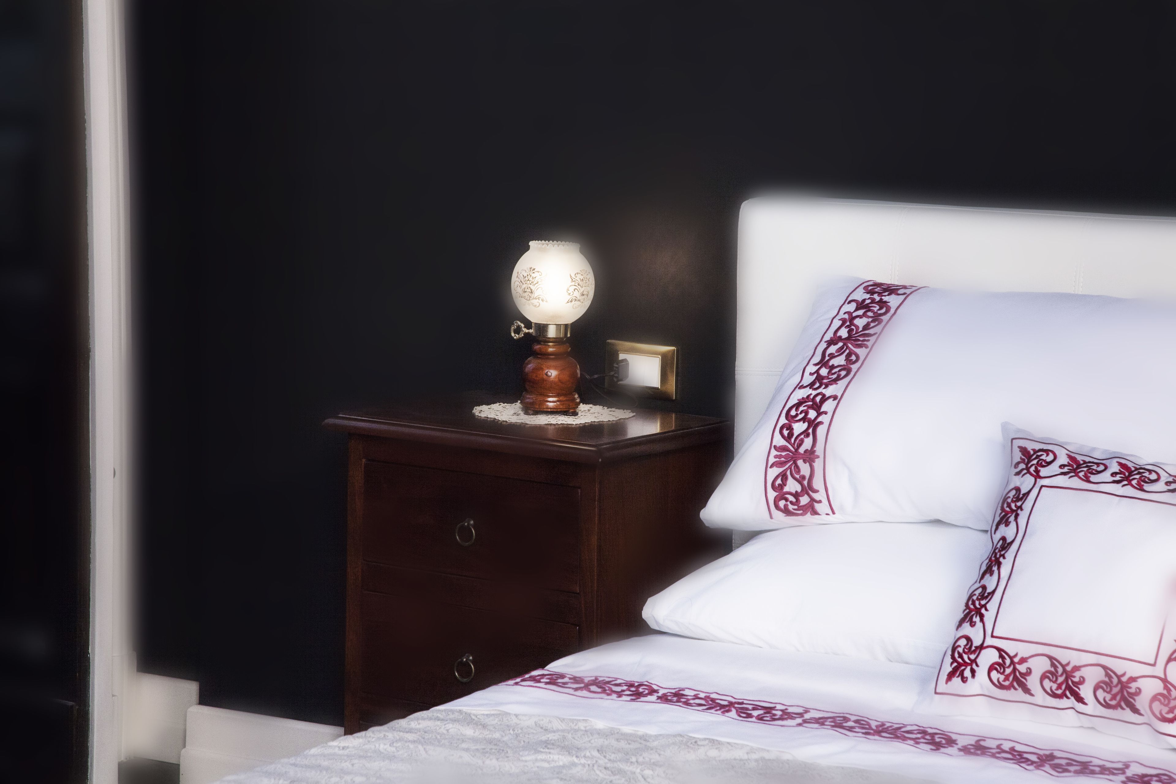 A neatly made bed with white bedding and decorative pillows, a wooden nightstand with a lamp, and a dark wall in the background.