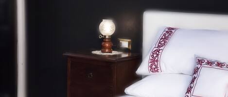 A neatly made bed with white bedding and decorative pillows, a wooden nightstand with a lamp, and a dark wall in the background.
