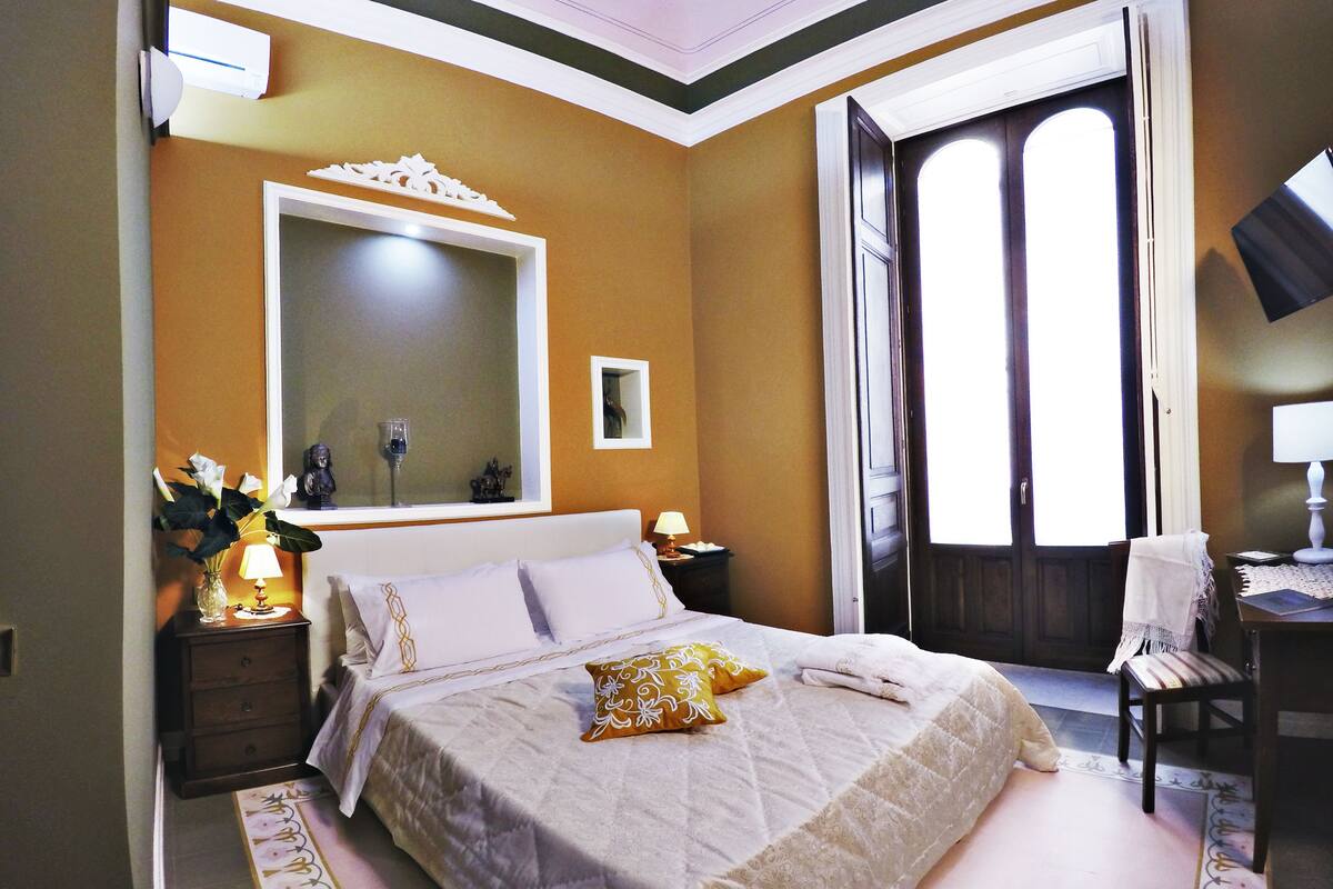 A bedroom with a large bed, a mirror, a nightstand with a lamp, and a window with curtains.