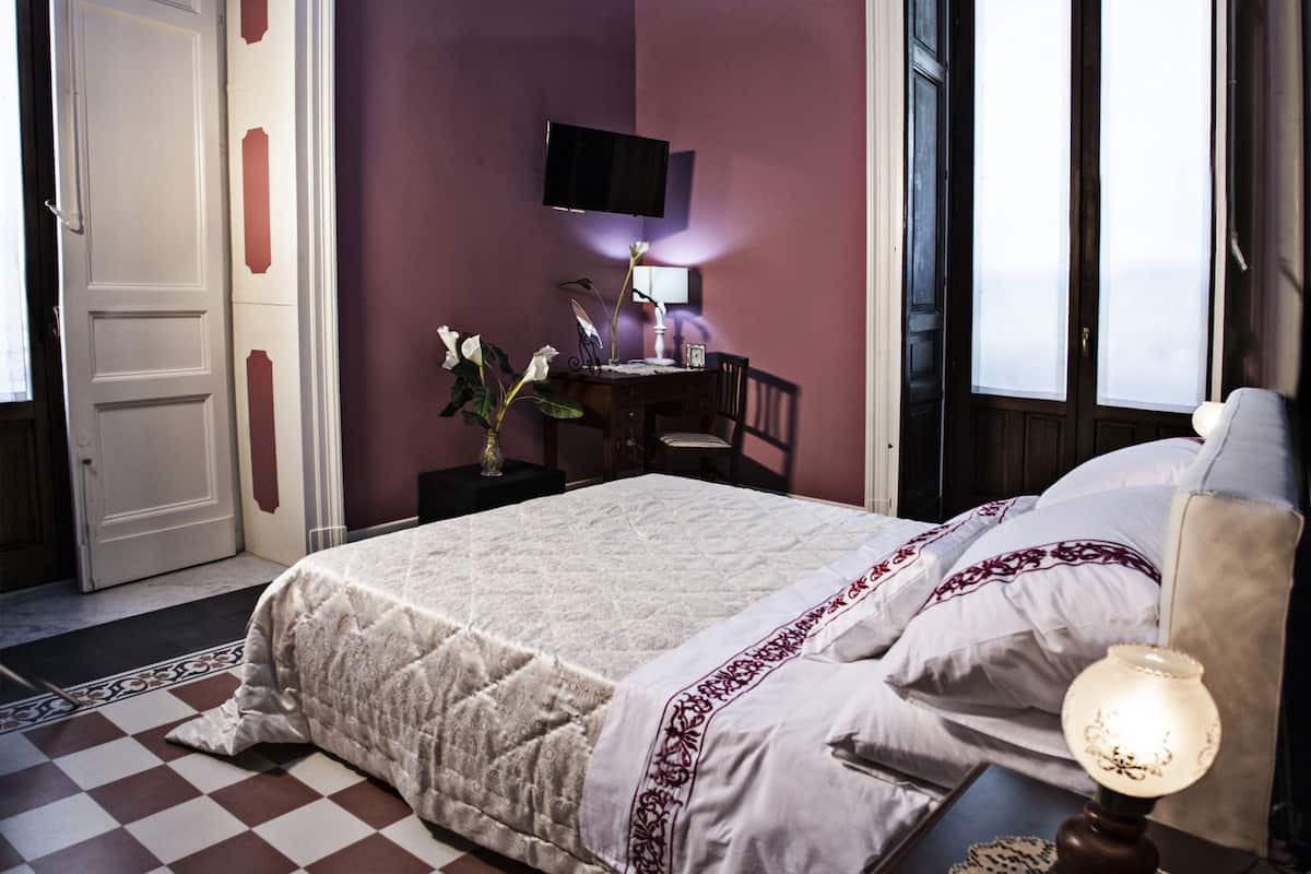 A bedroom with a checkered floor, a bed with a white and red bedspread, a nightstand with a lamp, a desk with a chair, and a TV mounted on the wall.