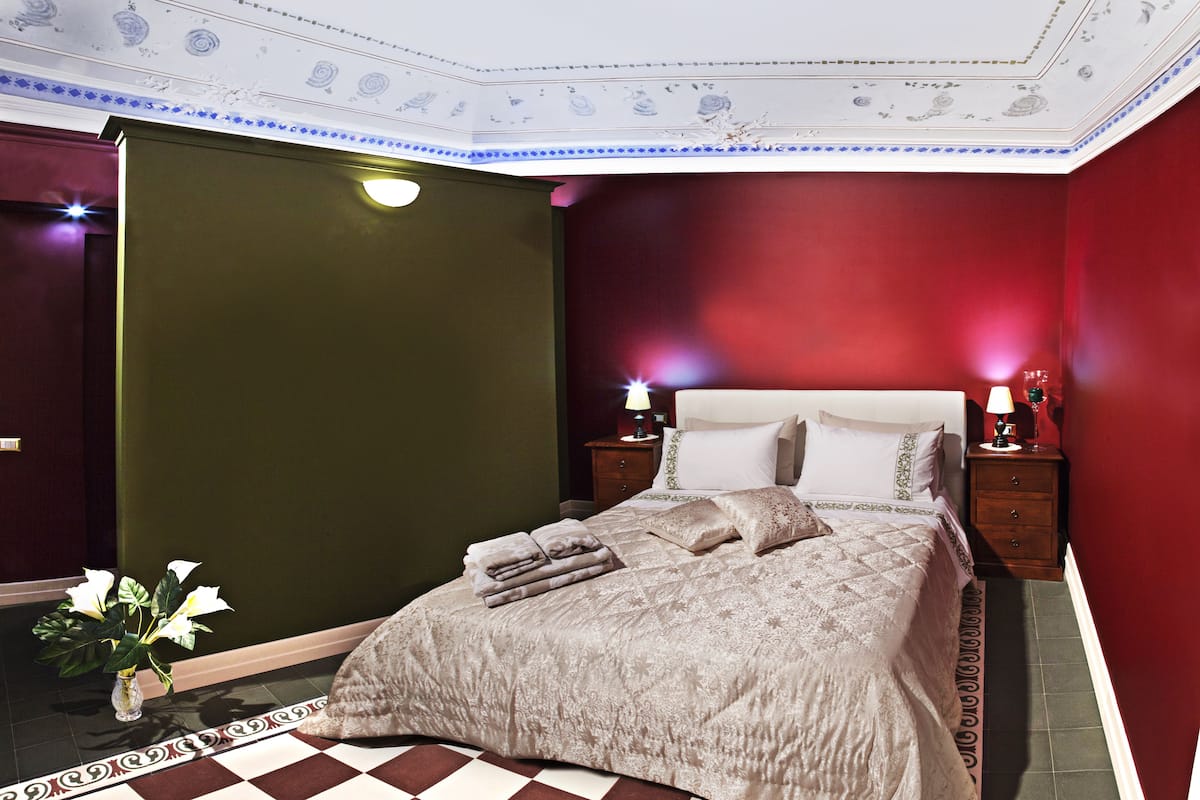 A bedroom with a checkered floor, a bed with a beige bedspread, two bedside tables with lamps, and a floral wall decoration.
