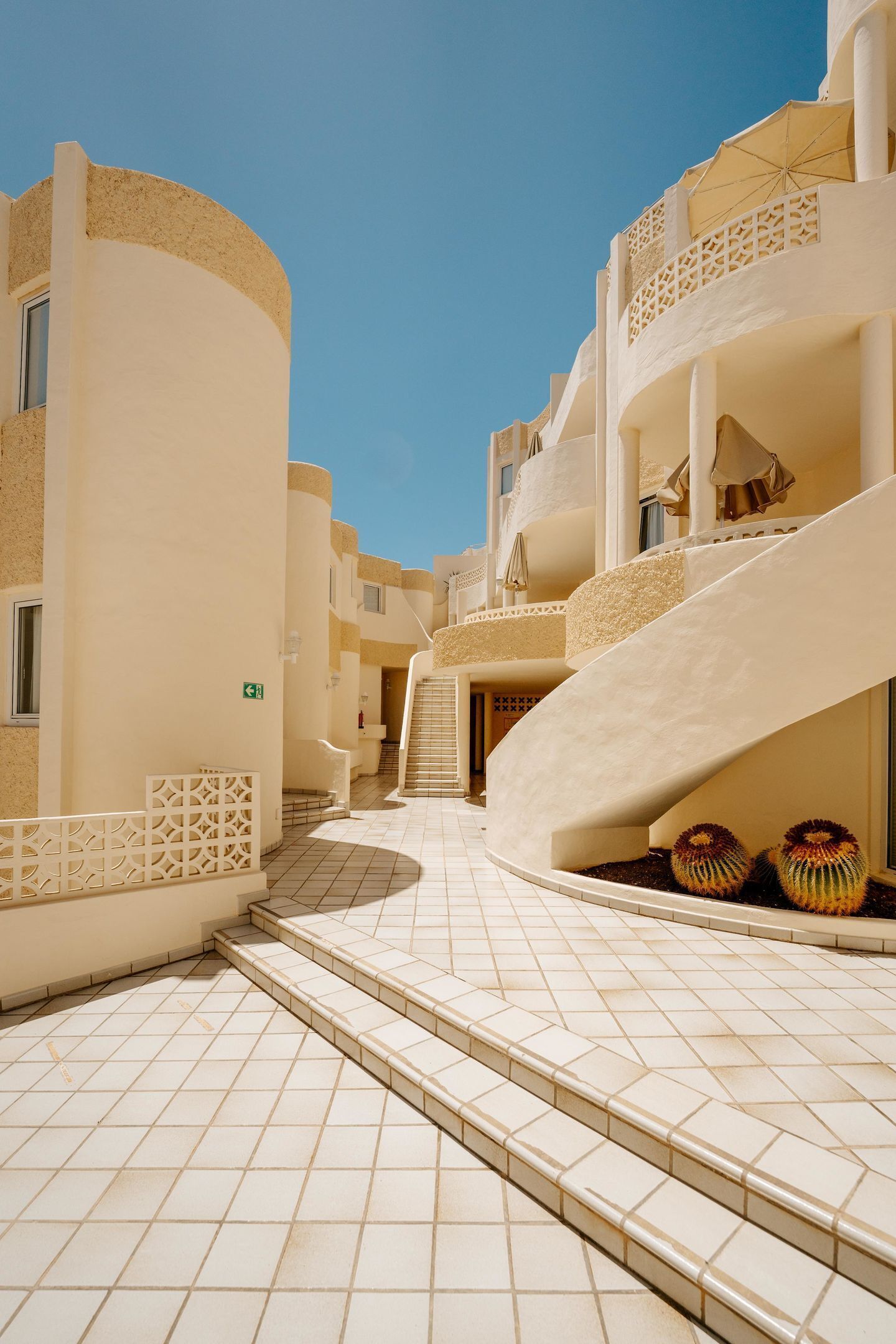 A hotel corridor with a curved staircase, tiled flooring, and a clear sky.
