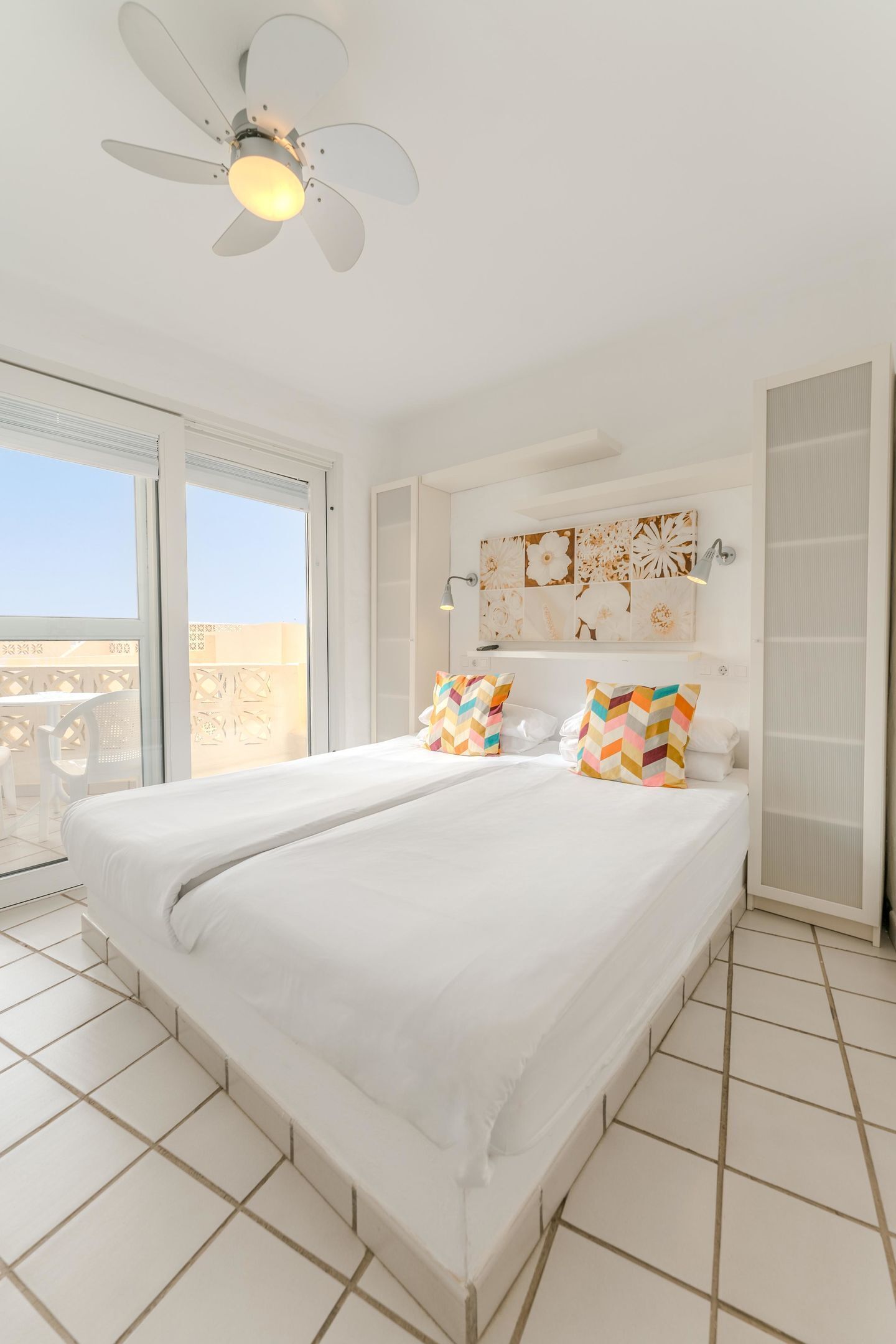 Ein Hotelzimmer mit einem großen Bett, einem Deckenventilator, einem Balkon mit einem weißen Stuhl und Blick auf die Stadt.