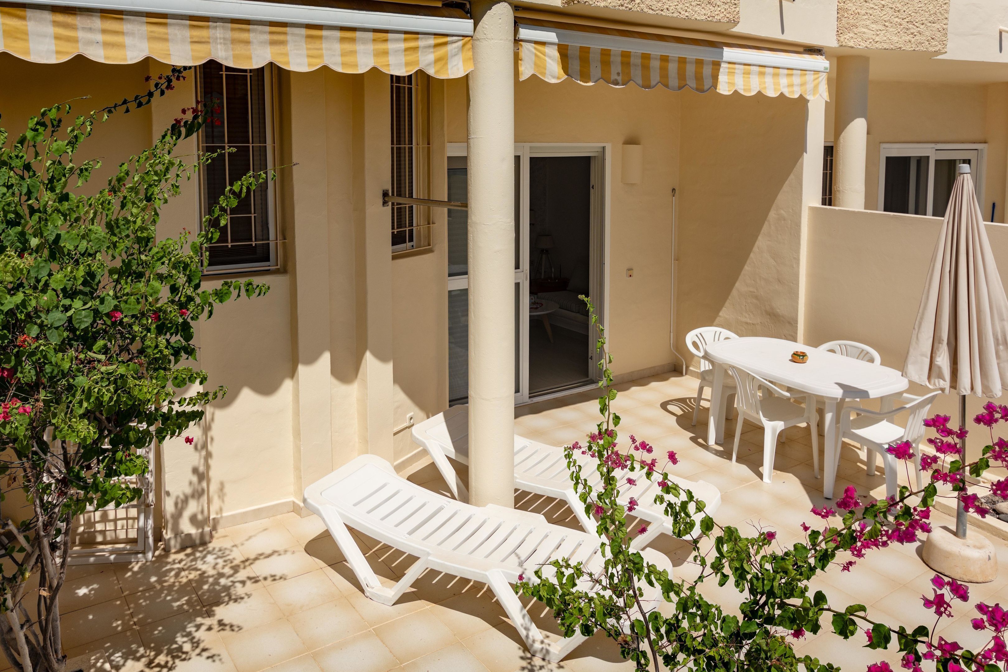 A sunlit patio with white lounge chairs, a table, and an umbrella.