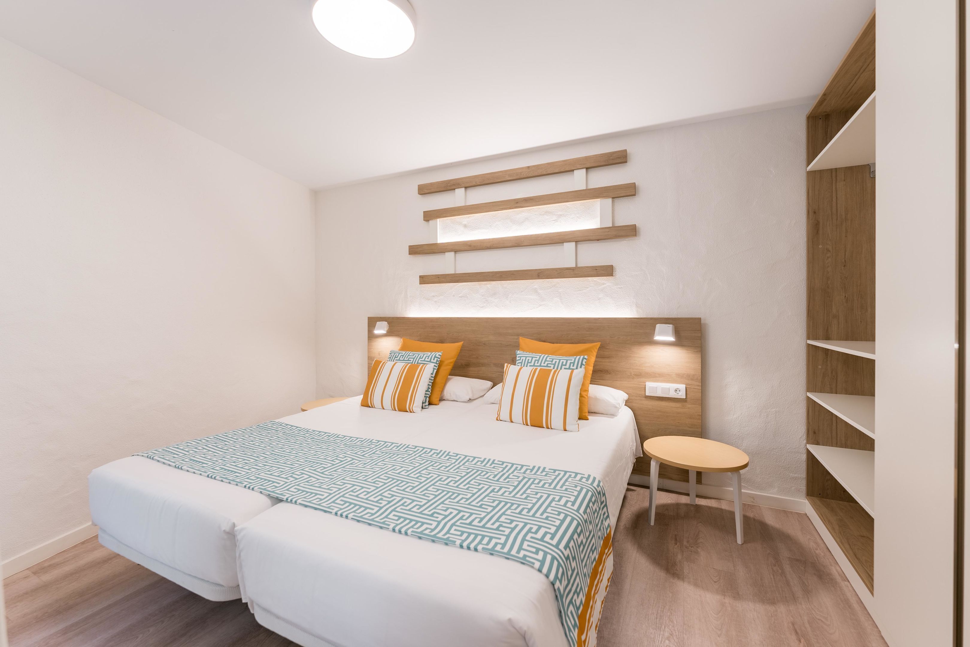 Une chambre d’hôtel moderne avec un grand lit, deux tables de chevet, un petit tabouret et des étagères en bois intégrées.
