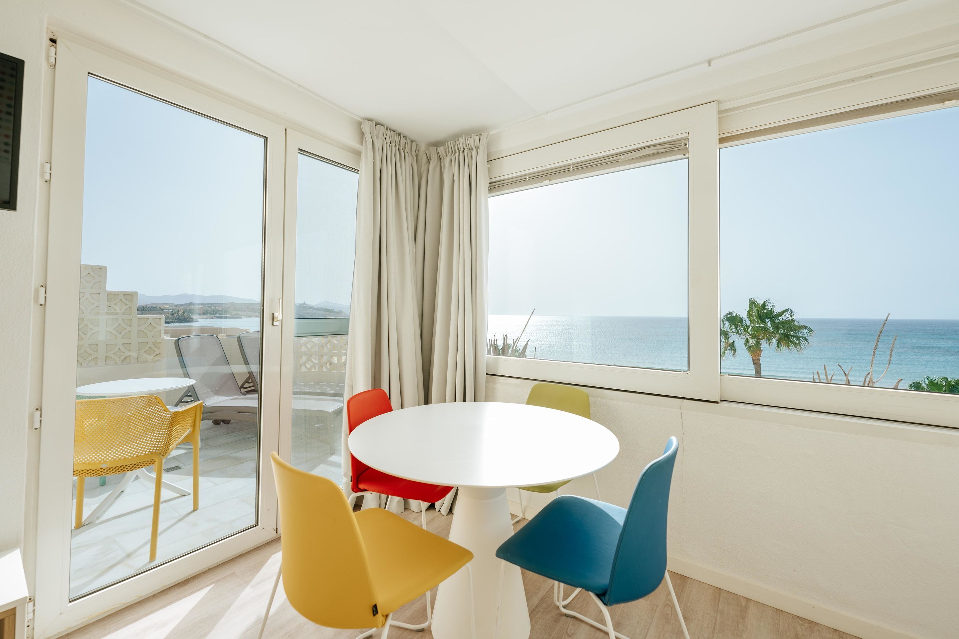Ein Balkon mit weißem Tisch und bunten Stühlen, mit Blick auf die Küste.