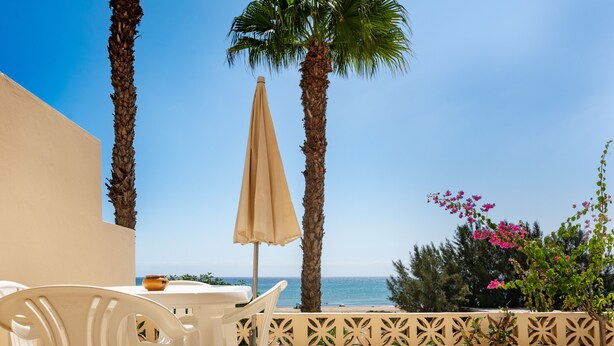 Una terraza con sillas de plástico blancas, una sombrilla color beige y vista al mar.