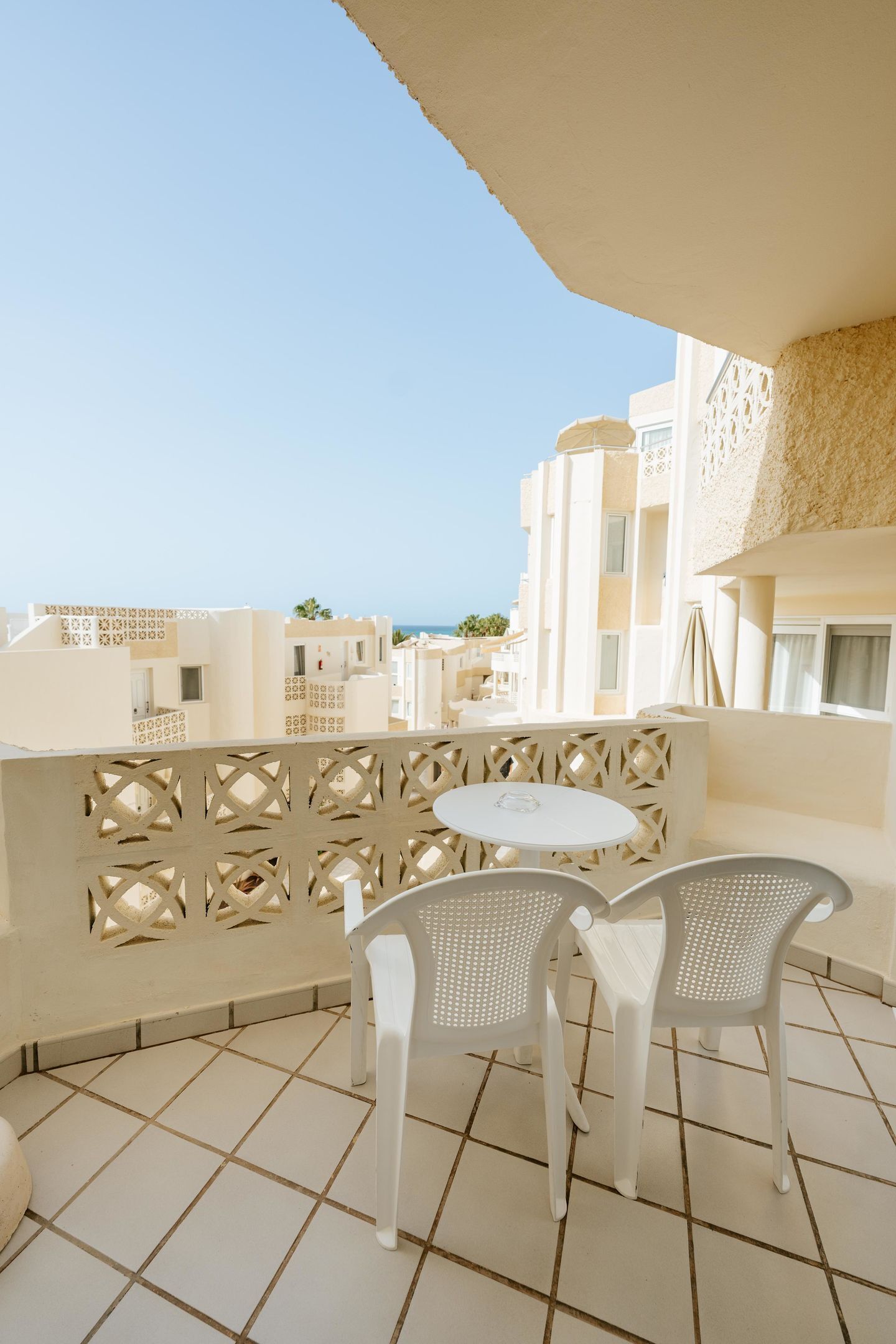A balcony with a white table and two chairs, overlooking a coastal area with buildings and the sea.