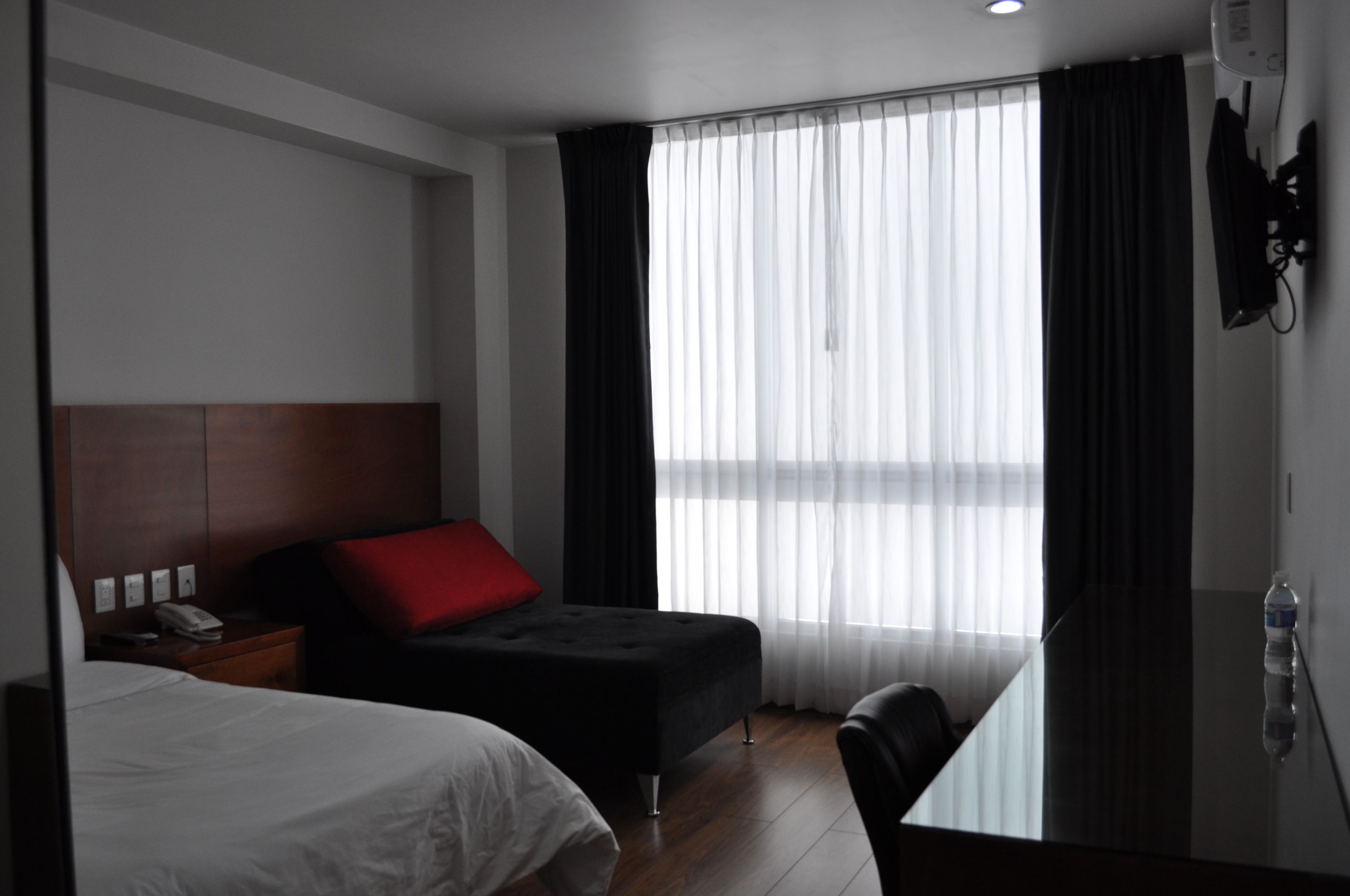 Quarto de hotel com cama, escrivaninha, cadeira, mesa de cabeceira e janela com cortinas.