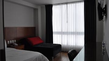 Quarto de hotel com cama, escrivaninha, cadeira, mesa de cabeceira e janela com cortinas.