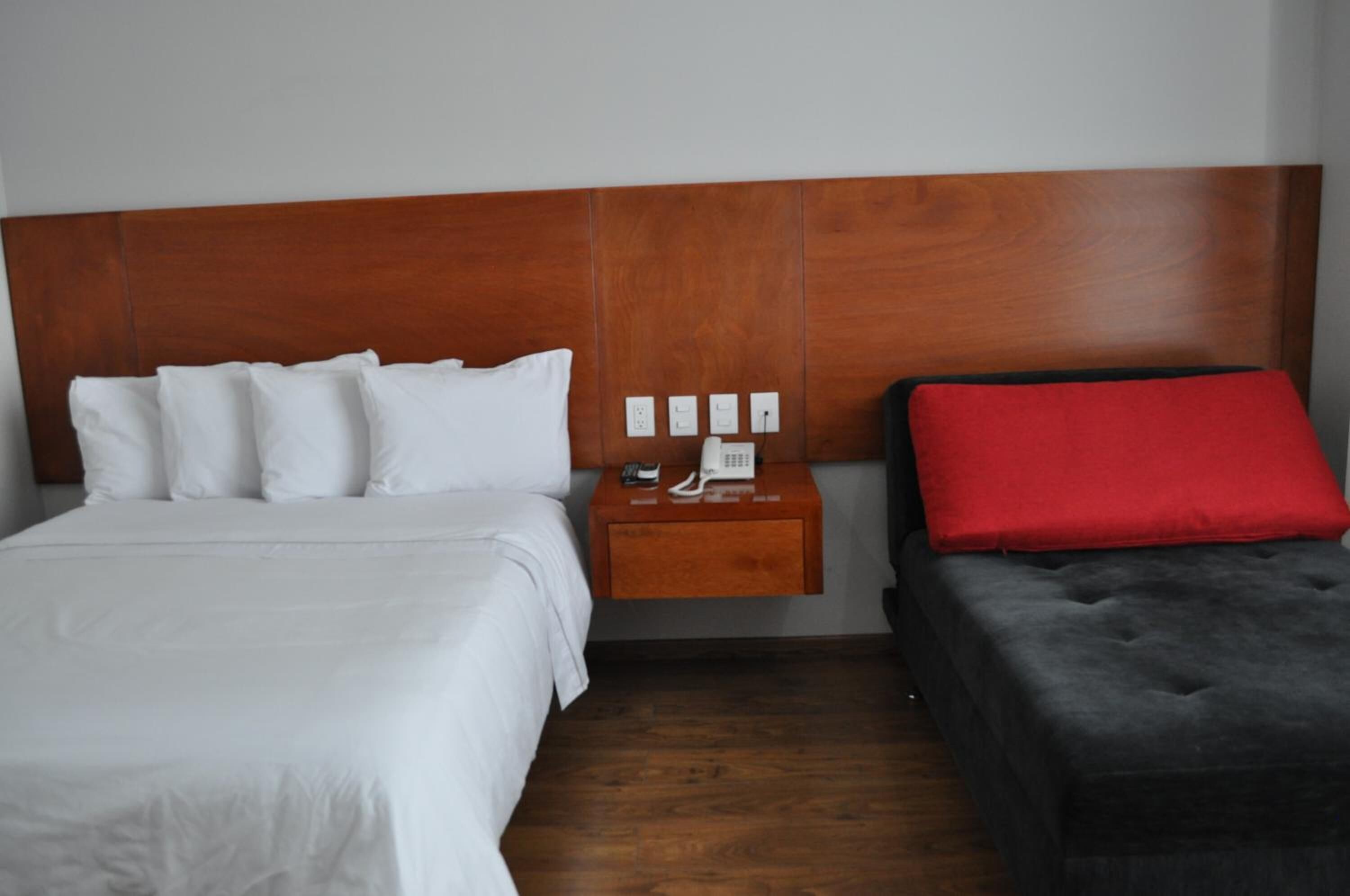 A hotel room with a wooden headboard, a bed with white bedding, a bedside table with a phone and a lamp, and a red pillow on a grey ottoman.