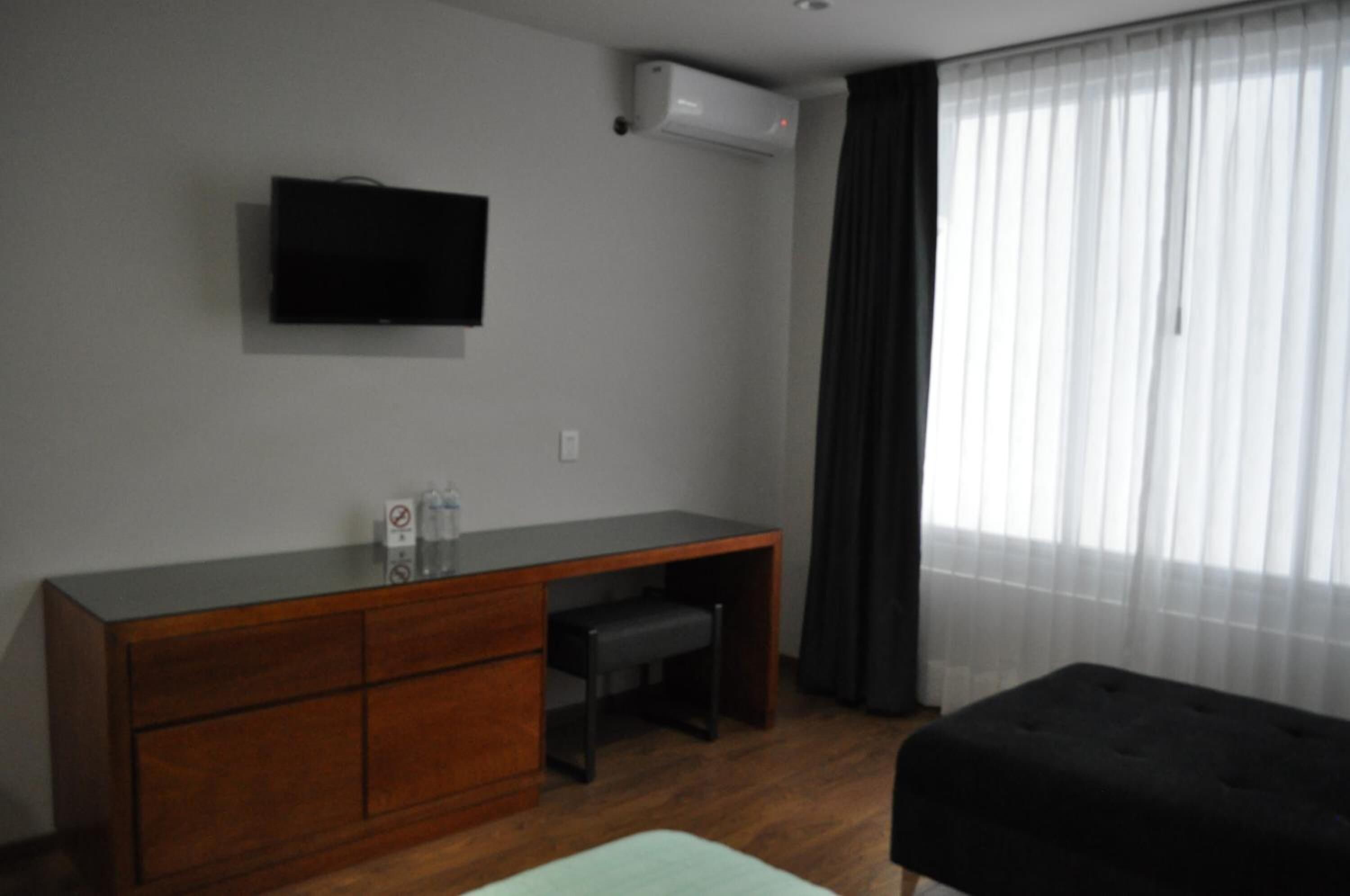 A room with a flat-screen TV mounted on the wall, a wooden desk with drawers, a bench, and a window with sheer curtains.