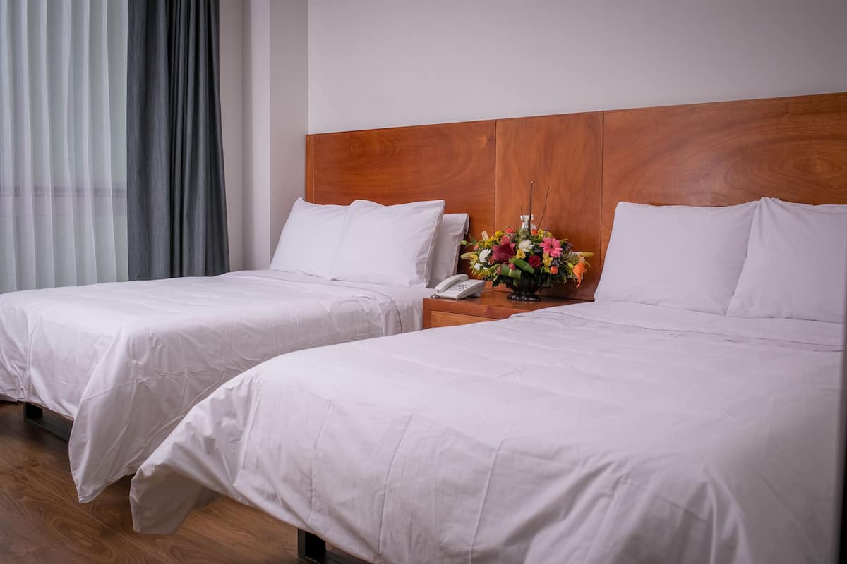 Une chambre d’hôtel avec deux lits, une tête de lit en bois, une table de chevet avec un téléphone et un vase de fleurs.